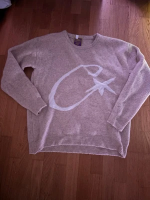 Beige stickad tröja med stjärnmotiv - Mysig beige stickad tröja  med ett stort vitt C och stjärna på framsidan. Tröjan har rund halsringning, långa ärmar och ribbstickade muddar. Perfekt för dig som gillar unika detaljer och en avslappnad stil.