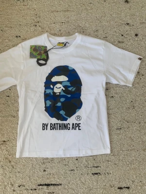 Vit BAPE t-shirt med blå camo print - Säljer en vit t-shirt från A Bathing Ape med det ikoniska BAPE-huvudet i blått camo-mönster på bröstet och texten 'BY BATHING APE' under. Klassisk rund hals och korta ärmar. Perfekt för dig som gillar streetwear och designer 