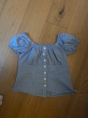 Rutig topp! - Söt blårutig blus med offshoulder och puffiga korta ärmar. Blusen har knappar framtill och resår vid axlarna för en snygg passform. Perfekt till jeans eller kjol för en trendig look.