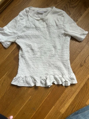 Vit topp med volang från Gina Tricot - Söt vit topp från Gina Tricot med korta ärmar och smockad kropp. Toppen har en volangkant nedtill som ger en extra detalj. Perfekt att styla med jeans eller kjol för en trendig look.