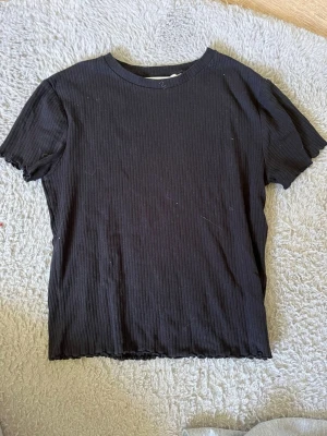 Svart ribbad t-shirt - En svart ribbad t-shirt med korta ärmar och rund hals. Den har små volanger på ärmarna. Storlek 170 men passar xs/s
