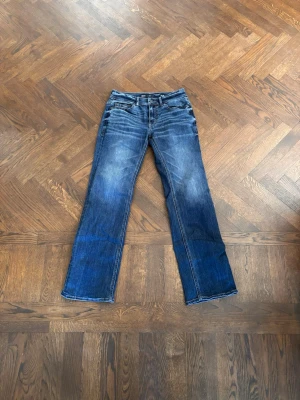 Blå bootcut jeans med slitningar - Säljer ett par blå bootcut jeans med snygga slitningar och tvättade detaljer. Jeansen har klassisk femficksdesign och normal midja. Materialet är denim i bomull och passformen är något utsvängd nedtill för en retro vibe.