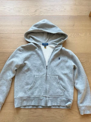 Grå hoodie från Polo Ralph Lauren - Snygg grå hoodie från Polo Ralph Lauren med dragkedja, två fickor framtill och blå broderad logga på bröstet. Insidan är fodrad med mjuk teddy för extra komfort. Klassisk huva och ribbade muddar vid ärmar och nederkant. Perfekt för en chill och avslappnad stil.
