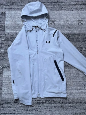  vindjacka från Under Armour - Snygg  vindjacka från Under Armour med svart logga på bröstet. Jackan har huva med dragsko, hel dragkedja framtill och svarta dragkedjor på fickorna. Tillverkad i ett lätt och smidigt material som passar perfekt för blåsiga dagar.