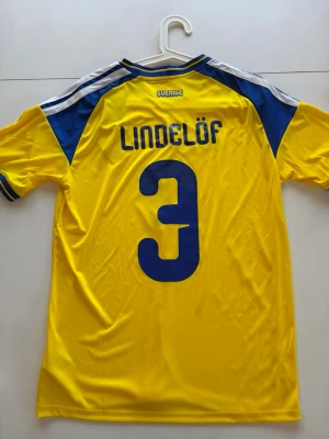 Sverige Lindelöf matchtröja gul - Gul matchtröja från Sveriges landslag med blå detaljer, Lindelöf och nummer 3 på ryggen. Tröjan har broderat landslagsemblem på bröstet och klassiska Adidas-ränder på axlarna. Tillverkad i lätt och ventilerande polyester, perfekt för fotboll.