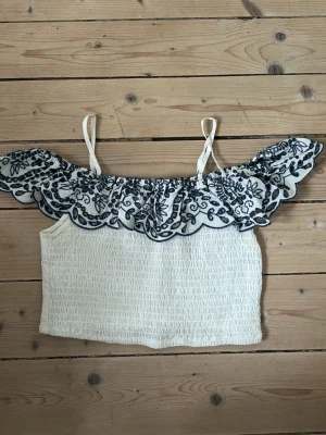 Vit broderad linnetopp Forever 21 - Supersöt vit linnetopp från Forever 21 med tunna axelband och volangkant upptill. Toppen har mörkblå broderade blommor längs volangen. 