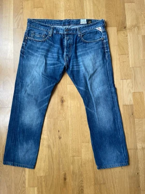 Blå raka jeans från Replay - Snygga blå jeans från Replay med raka ben och klassisk femficksdesign. Jeansen har en tydlig tvättad look med slitningar och knäppning med knappar. Perfekta för en avslappnad och trendig stil.