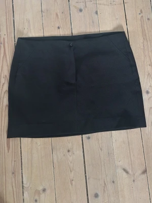 Svart kort kostymkjol - En stilren svart kortkjol i kostymmaterial med knappstängning i midjan. Kjolen har en enkel, rak passform och är perfekt att matcha med olika toppar. 
