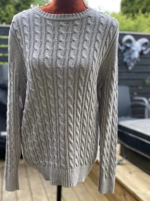 Ljusgrå kabelstickad tröja L - Tröja med rund halsringning och långa ärmar. Tröjan har ett flätstickat mönster över hela plagget, vilket ger en klassisk och texturerad look. Muddar finns vid ärmslut och nederkant. Längd: 71 cm Bystvidd: 53 cm  Inga bilder på mig! Katter finns i hemmet!
