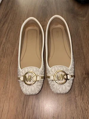 Beige ballerinaskor Michael Kors - Snygga ballerinaskor från Michael Kors i beige med klassiskt MK-mönster och guldfärgade detaljer framtill. Skorna har rund tå, låg klack och vita kanter. Tillverkade i textil med syntetdetaljer och en stor MK-logga i metall på tån. Har används 1 gång har en liten skada på höger sida 