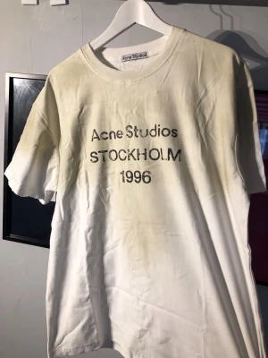 Vit Acne Studios t-shirt med tryck - Vit t-shirt från Acne Studios med svart texttryck på bröstet där det står 'Acne Studios STOCKHOLM 1996'. Klassisk rund hals och korta ärmar. Tillverkad i mjuk bomull för en skön och avslappnad look.