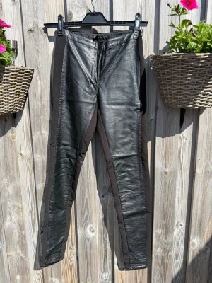 Svarta leggings i fuskskinn från H&M (34) - Säljer ett par svarta leggings från H&M i storlek 34. Framsidan är i glansigt fuskskinn och sidorna samt sidorna är i stretchigt tyg för en snygg och bekväm passform. Hög midja och smala ben ger en trendig look.