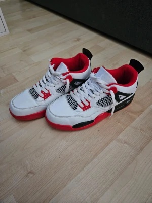 Air Jordan 4 Retro White/Red/Black - Säljer ett par Air Jordan 4 Retro sneakers med vit läderöverdel, röda och svarta detaljer samt meshpaneler på sidorna. Skorna har en klassisk siluett med röda insidor och vita skosnören. Perfekt för dig som gillar ikoniska sneakers och streetstyle kan diskutera om priset.