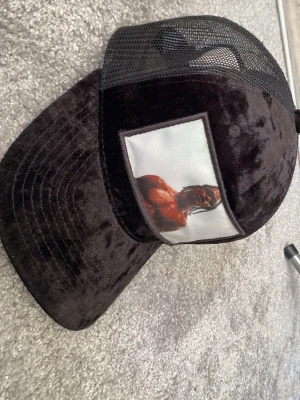 Svart meshkeps med Travis scott på - Säljer en svart keps med mesh baktill och sammetsliknande material framtill. På framsidan finns ett stort tygmärke med ett färgstarkt tryck av en person. Kepsen har böjd skärm och justerbar passform bak. Pris kan diskuteras 