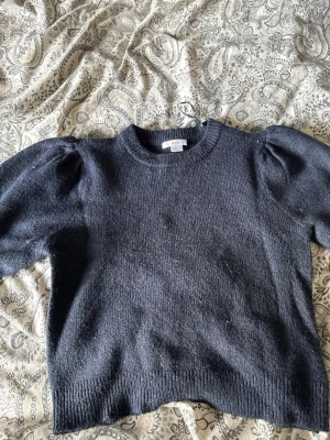 Mörkblå stickad tröja med puffärm - Mörkblå stickad tröja med rund hals och puffade axlar. Tröjan har ribbstickade muddar vid ärmslut och nederkant, vilket ger en snygg och bekväm passform. Perfekt för dig som gillar klassiska och trendiga detaljer.