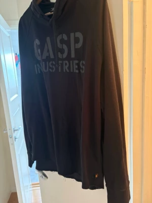 Svart hoodie från GASP Industries - Cool svart hoodie från GASP Industries med stor logga på bröstet. Hoodien har lång ärm och klassisk huva, perfekt för dig som gillar en sportig och avslappnad stil. Materialet är mjukt och bekvämt. 