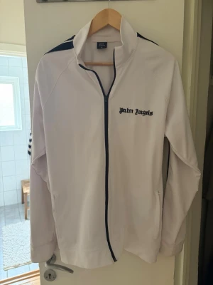 Vit ziptröja från Palm Angels - Säljer en vit ziptröja från Palm Angels med svarta detaljer längs dragkedjan och axlarna. Tröjan har hög krage, två fickor och logga broderad på bröstet. Perfekt för en sportig och trendig look.