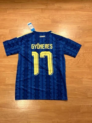 Blå Sverige fotbollströja Gyökeres - Säljer en blå Sverige fotbollströja från Adidas med Gyökeres och nummer 17 i gult på ryggen. Tröjan har korta ärmar, gul krage och gula ränder på axlarna samt Sveriges landslagsemblem på bröstet. Tillverkad i lätt och ventilerande material.