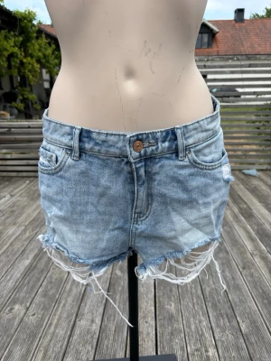 Lågmidjade jeansshorts med fransar  - Säljer ett par ljusblå denimshorts med råa, fransiga kanter och slitningar framtill. Klassisk femficksmodell med knapp och dragkedja. Perfekta för varma dagar och ger en avslappnad vibe.