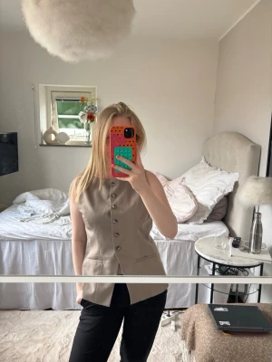Beige väst med knappar - Stilren beige väst med asymmetrisk knäppning framtill och dekorativa metallknappar. Västen har två fickor och är ärmlös med rak passform. Perfekt att styla över en skjorta eller t-shirt för en trendig look.