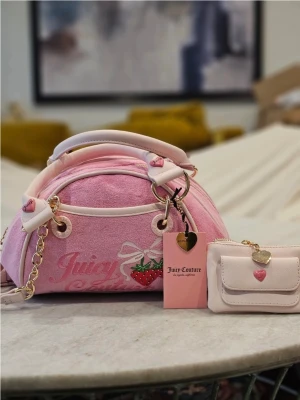 Rosa handväska Juicy Couture - Söt rosa handväska från Juicy Couture i mjuk velour med vita detaljer och guldfärgade kedjor. Väskan har ett jordgubbsmotiv, hjärtformade detaljer och matchande liten plånbok. Perfekt för dig som gillar gulliga accessoarer och vill sticka ut.
