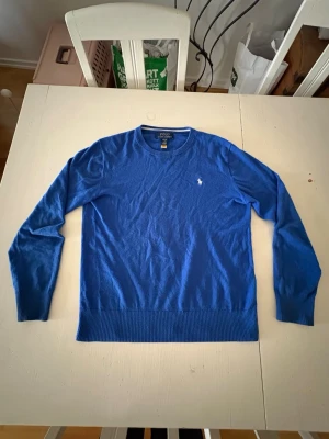 Blå tröja från Polo Ralph Lauren - Snygg blå tröja från Polo Ralph Lauren med rund halsringning och den klassiska broderade loggan på bröstet. Tröjan har långa ärmar och ribbade muddar vid ärmslut och nederkant. Perfekt för en clean och stilren look.