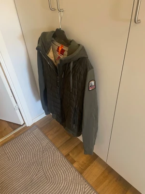 Grön quiltad jacka med huva från Parajumper  - Snygg grön jacka från Primark med quiltad front, huva och dragkedja. Jackan har ribbade muddar, två sidofickor och en bröstficka med dragkedja. Ärmarna är släta och jackan har en patch på ena ärmen. Perfekt för kyliga dagar.