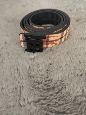 Burberry bälte  - Snyggt bälte från Burberry med klassiskt rutigt mönster i beige och i svart den är tvåsidig. Bältet har ett svart metallspänne med Burberrys logotyp. Perfekt accessoar för att lyfta din outfit. 