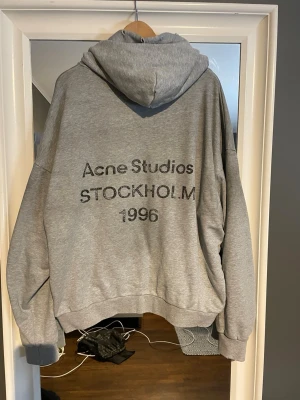 Acne Studios 1996 hoodie  - En grå Acne Studios hoodie med ”Stockholm 1996” tryck på ryggen. Storleken är M och den är gjort av 100% bomull. Köpt på Vinted. Slitningar på luvan och muddarna är design. Sparsamt använd med lappen kvar (se bild nr 2 och 3). Priset är inte hugget i sten, skriv gärna om du är intresserad!