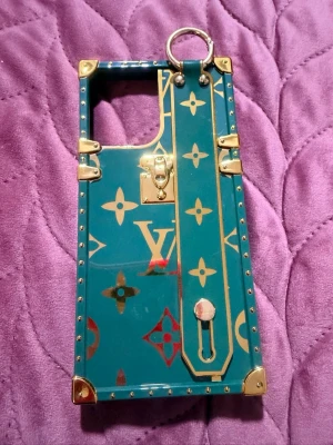  Mobilskal med handrem för iphone 12 pro max - Snyggt mobilskal i blått med guldfärgade detaljer och LV-inspirerat mönster. Skalets form är kantig med metallhörn och nitar, och det har en praktisk handrem med ring för extra grepp. Materialet är hårdplast med metalliska inslag för en lyxig känsla.