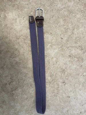 Blått flätat bälte med läderdetaljer - Snyggt blått flätat bälte med vita inslag och bruna läderdetaljer vid spännet. Bältet har ett klassiskt metallspänne och passar perfekt till jeans eller chinos. Ger en stilren touch till din outfit.