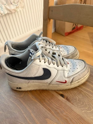 Nike Air Force 1 vita sneakers - Nike Air Force 1 sneakers i vitt läder med svart swoosh på sidan och små röda detaljer. Klassisk låg modell med perforerad tå och vita skosnören. Yttersulan är ljus och har AIR-tryck på sidan. Perfekt för dig som gillar streetstyle.