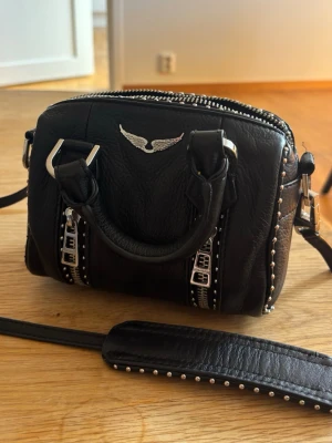 Svart handväska Zadig & Voltaire - Cool svart handväska i läder från Zadig & Voltaire med silverfärgade nitar, detaljerade spännen och en ikonisk vinge framtill. Väskan har både handtag och justerbar axelrem med nitar, samt dragkedja upptill. Perfekt för dig som gillar edgy och trendig stil.