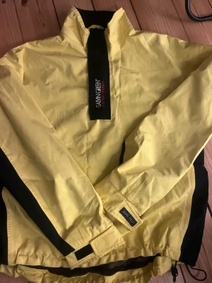 Golfjacka från Galvin Green - En gul vindjacka från Galvin Green med svarta detaljer och hög krage mandatory . Jackan har just neterbara muddar och midja med kardbor dren , samt dragkedja och kardborreknäppning framtill. Tillverkad i ett lätt och vindtätt Gore tex material 