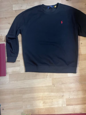  Polo Ralph Lauren swetshirt  - Klassisk svart sweatshirt från Polo Ralph Lauren med rund halsringning och den ikoniska röda logon broderad på bröstet. Tröjan har långa ärmar och ribbade muddar vid ärmslut och nederkant. Perfekt för en avslappnad och stilren look.
