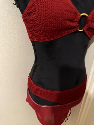 Röd bikini med meshkjol och ringdetalj - Snygg röd bikini med ribbad struktur och guldfärgad ringdetalj framtill på toppen. Matchande meshkjol med dragsko på sidan för extra stilpoäng. Perfekt för strandhäng eller poolparty.