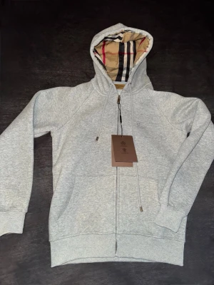 Grå hoodie från Burberry med dragkedja - Snygg grå hoodie från Burberry med dragkedja och klassiskt rutigt foder i huvan. Hoodien har fickor framtill, justerbar dragsko och diskret Burberry-logga på bröstet. Tillverkad i mjukt bomullsmaterial för maximal komfort.