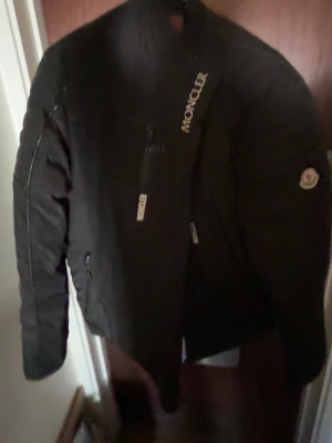 Svart dunjacka från Moncler - Snygg svart dunjacka från Moncler med diskret logga på ärmen och dragkedja framtill. Jackan har hög krage och en bröstficka med dragkedja. Perfekt för kalla vinterdagar och riktigt stilren look.