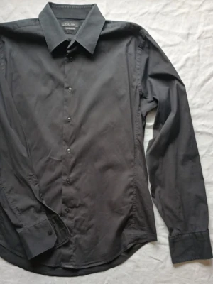 Svart skjorta med lång ärm - Klassisk svart skjorta från Selected Homme med lång ärm och knappar framtill. Skjortan har en stilren krage och är tillverkad i ett mjukt bomullsmaterial. Perfekt för dig som gillar en enkel och clean look.