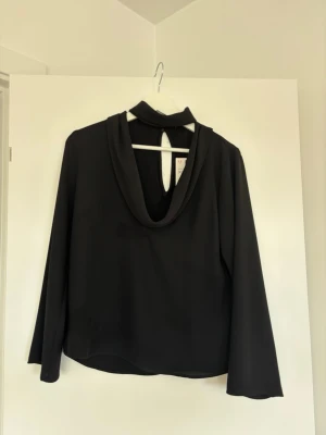 Svart blus med draperad krage - Stilren svart blus från Zara med långärmad design och draperad krage. Blusen har en snygg öppning framtill med en knappstängning i nacken. Tillverkad i ett mjukt, lätt tyg som ger ett elegant fall.
