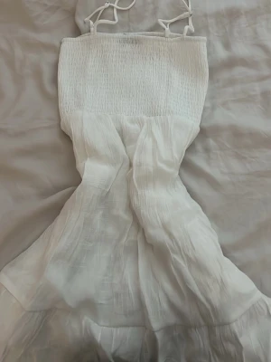 Vit sommarklänning med smock - Luftig vit klänning med smala axelband och smockad överdel. Kjolen är lätt utsvängd med volangdetaljer och tillverkad i ett tunt, mjukt tyg som passar perfekt för varma dagar. Enkel och stilren design som ger en fräsch look.