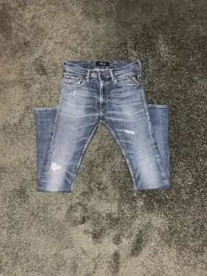 Replay blå jeans med slitningar - Säljer ett par snygga Replay jeans i blå tvätt med coola slitningar och detaljer på både fram- och baksida. Klassisk femficksmodell med dragkedja och knapp, samt Replay-logga på bakfickan. Jeansen har en slim-fit passform och är gjorda i jeansmaterial. Har du frågor är det bara att skicka! Priset är inte hugget i sten – kom med ett rimligt bud.