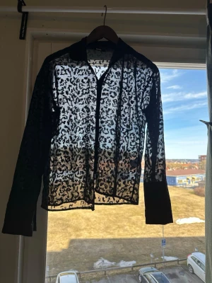 Svart transparent leopardmönstrad skjorta - Snygg svart skjorta med transparent tyg och leopardmönster. Skjortan har klassisk krage och långa ärmar, perfekt för dig som vill sticka ut med en trendig look. Passar till många olika outfits och ger en edgy vibe.