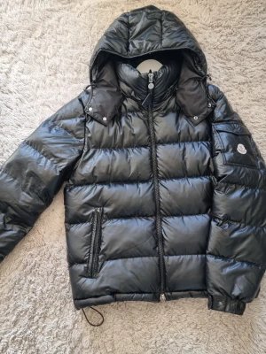 Svart dunjacka från Moncler - Snygg svart dunjacka från Moncler med glansig finish, huva och dragkedja framtill. Jackan har två sidofickor med dragkedja, Moncler-logga på ärmen och justerbara muddar. Perfekt för kalla vinterdagar och riktigt trendig look.