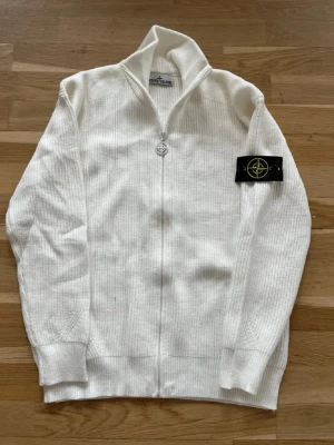  Stone Island cardigan  - OBS: SJÄRNAN PÅ KEDJAN FINNS INTE KVAR, och den är lite mer vitare på bild . Säljer en vit stickad ziptröja från Stone Island med ribbad struktur och ikonisk svart patch med gult kompassmärke på ärmen. Tröjan har hög krage, dragkedja framtill och långa ärmar med breda muddar. Perfekt för dig som gillar streetwear och snygga detaljer.