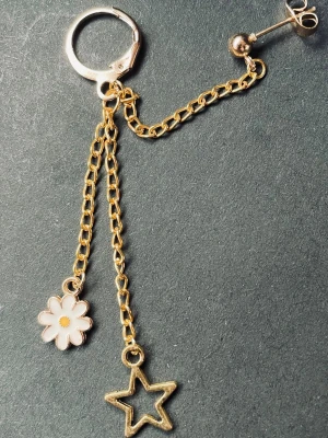 By prisförslag- 1 st Guldörhänge med kedjor, blomma & stjärna - Unikt örhänge i guldfärgad metall med två kedjor, en med en vit emaljerad blomma och en med en öppen stjärnberlock. Örhänget har både ring och plupp för olika stilar. Perfekt för dig som gillar lekfulla detaljer och trendig design.
