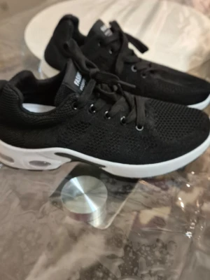 Svarta mesh sneakers ORTHOdaily - Snygga svarta sneakers från ORTHOdaily med ovandel i mesh-tyg och vita, tjocka sulor. Skorna har svarta skosnören och en sportig look med textdetaljer på plösen. Perfekta för dig som gillar stilrena och bekväma sneakers.