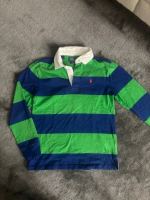 Randig rugbytröja från Polo Ralph Lauren för barn  - Klassisk långärmad rugbytröja från Polo Ralph Lauren med breda blå och gröna ränder, vit krage och knappslå. Ikonisk röd logga broderad på bröstet. Perfekt för en sportig och avslappnad stil. Tröjan passar barn M 