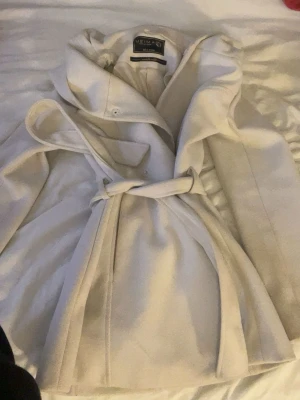 Ljusbeige kappa från Wilma - Stilren ljusbeige kappa från Wilma med knytskärp i midjan och bred krage. Kappan har en klassisk, rak passform och är tillverkad i ett mjukt material som ger en elegant look. Perfekt för dig som vill ha en tidlös och snygg ytterjacka.