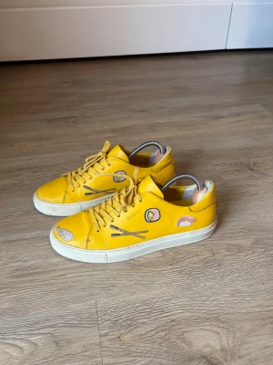 Axel Arigato Sushi Sneaker - Unika gula sneakers från Axel Arigato med broderade sushi- och ätpinnemotiv på sidorna. Skorna har vita sulor, gula skosnören och är tillverkade i läder. Perfekta för dig som vill sticka ut med färg och coola detaljer. Över lag i väldigt bra skick. Kolla gärna in mina andra annonser också